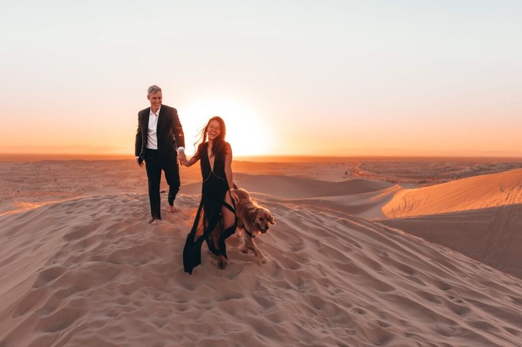 Full body of positive laughing couple holding hands and running with Golden Retriever dog on sandy ground of desert in sunlight